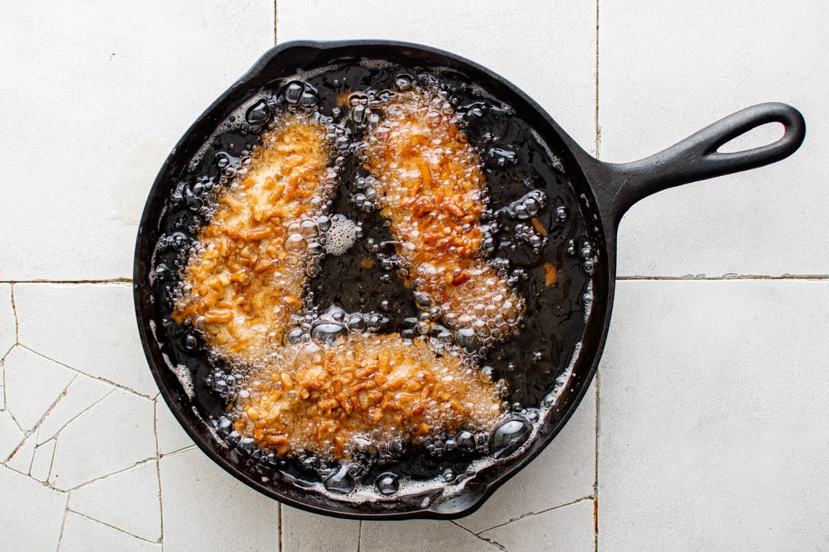 3 pretzel chicken tenders frying in oil in a skillet.