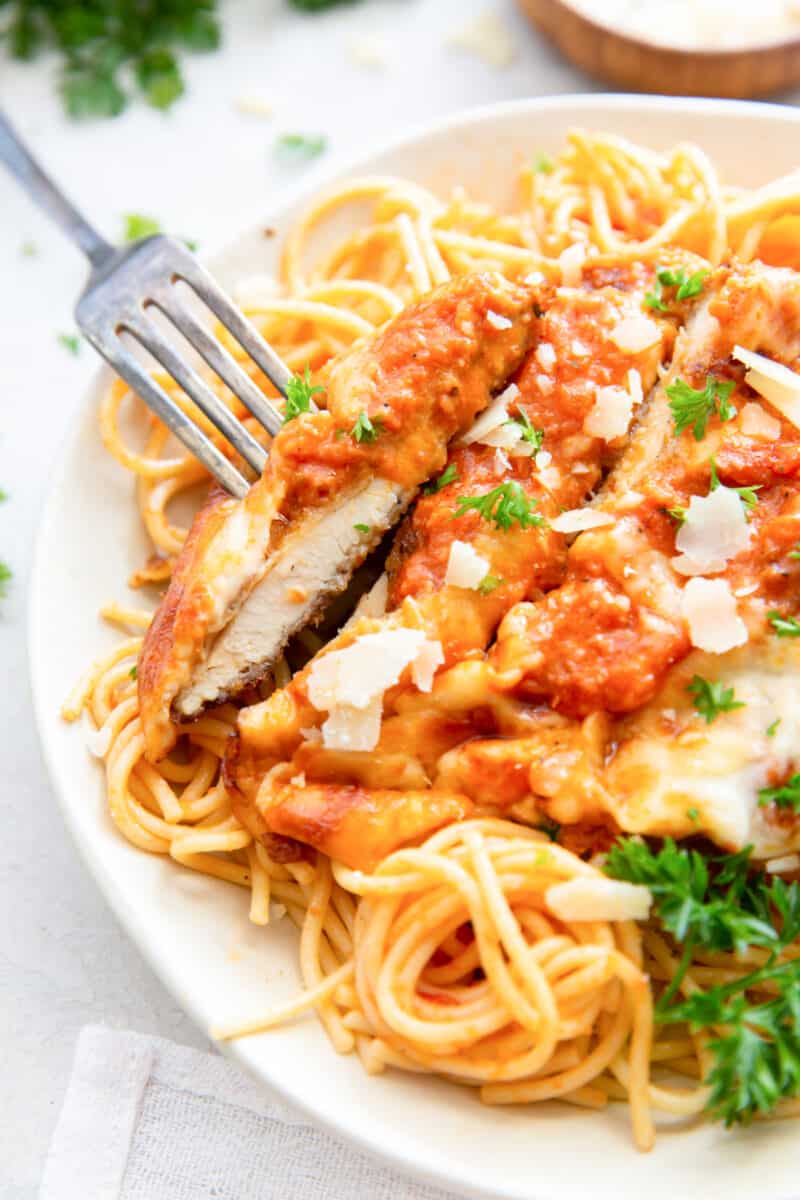 fork with a slice of chicken parmesan on top of spaghetti on a white plate