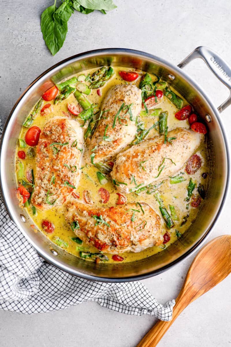 overhead image of chicken breasts in a skillet with pesto cream sauce, tomatoes, and asparagus