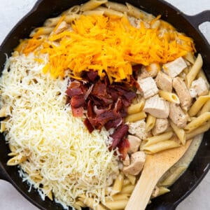 shredded cheese and bacon added to crack chicken pasta in a skillet with a wood spoon