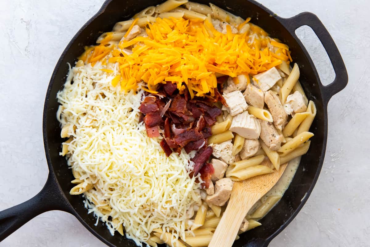 Shredded cheese and bacon added to crack chicken pasta in a skillet with a wood spoon.