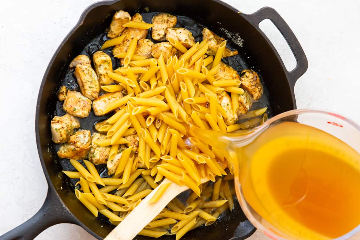 Pouring chicken broth into a skillet with penne noodles and chicken.