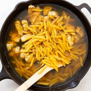 penne pasta and chicken broth added to skillet with chicken with a wood spoon