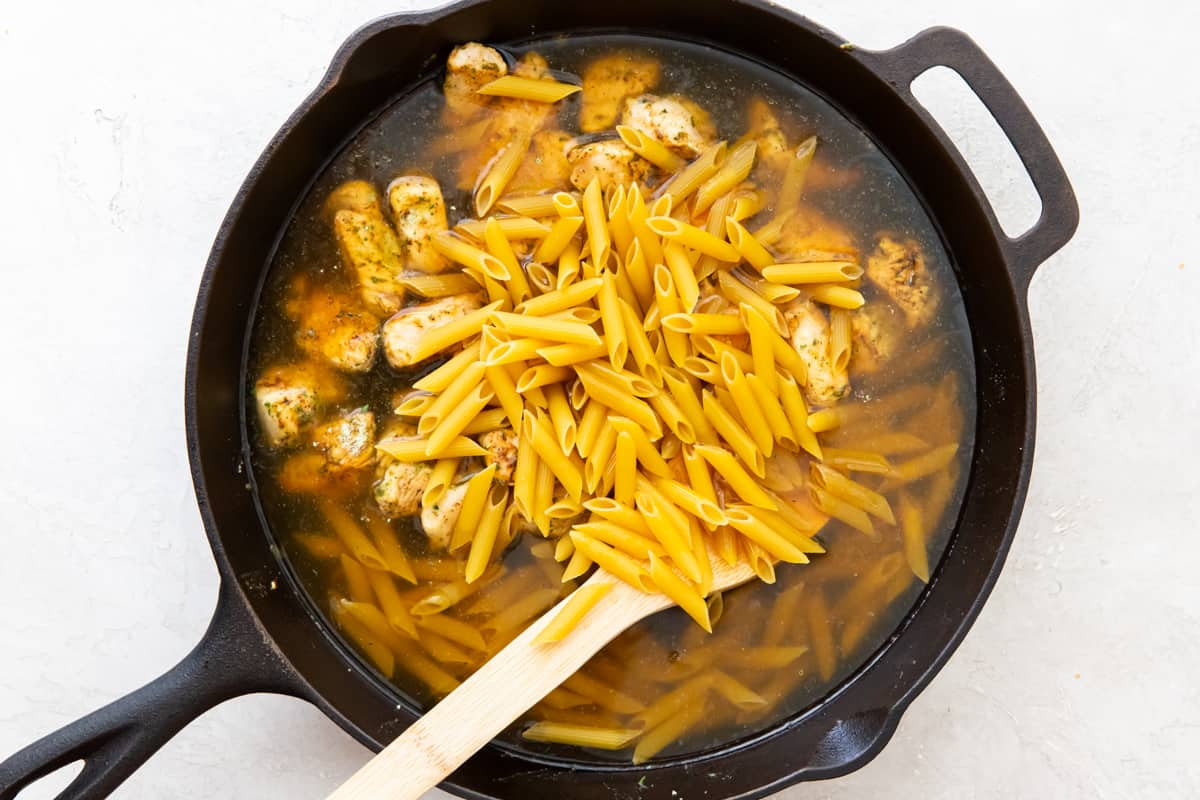 Penne pasta and chicken broth added to skillet with chicken with a wood spoon.