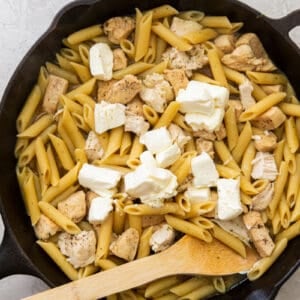 cream cheese cubes added to pasta and chicken in a skillet with a wood spoon