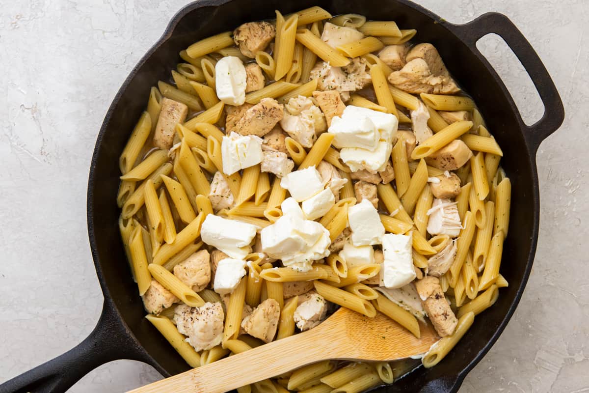 Cream cheese cubes added to pasta and chicken in a skillet with a wood spoon.