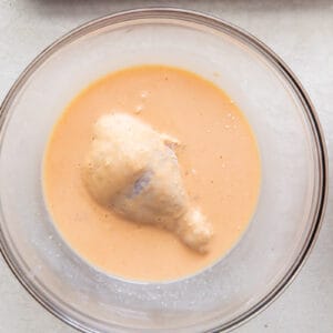 dipping a piece of flour coated chicken in a glass bowl of marinade
