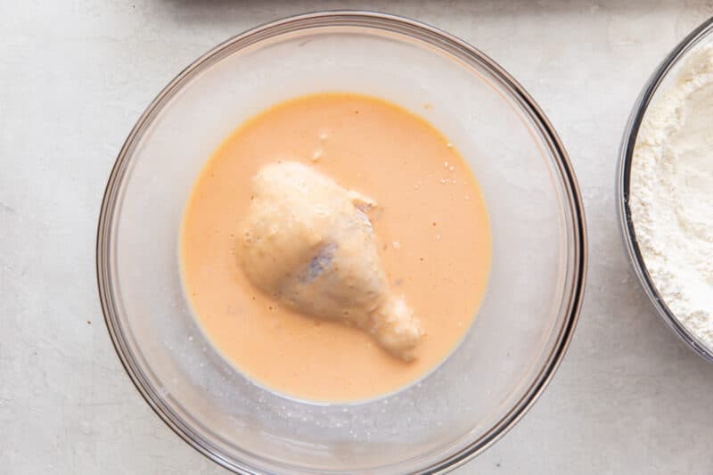 dipping a piece of flour coated chicken in a glass bowl of marinade