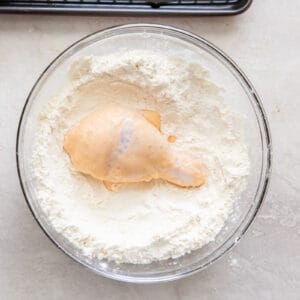 dredging a piece of marinated chicken in flour in a glass bowl