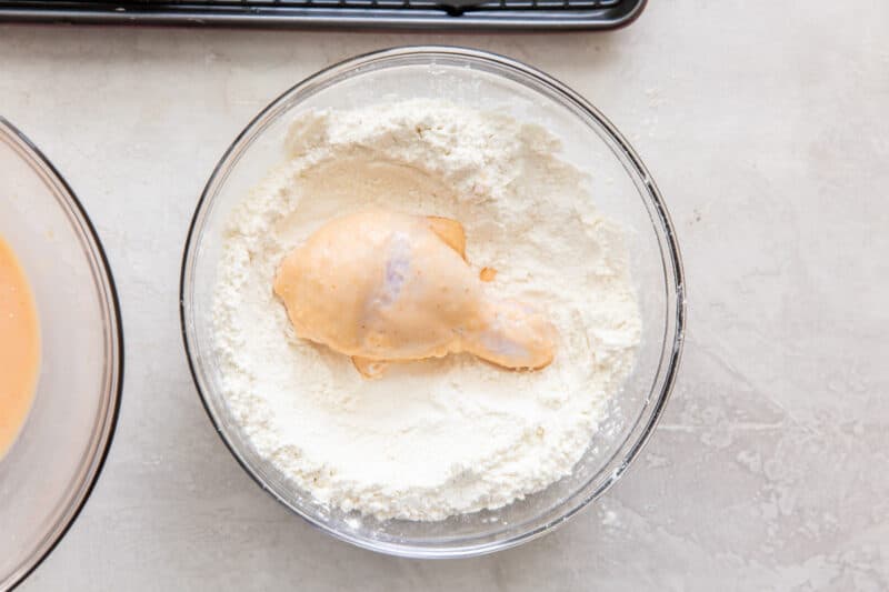 dredging a piece of marinated chicken in flour in a glass bowl