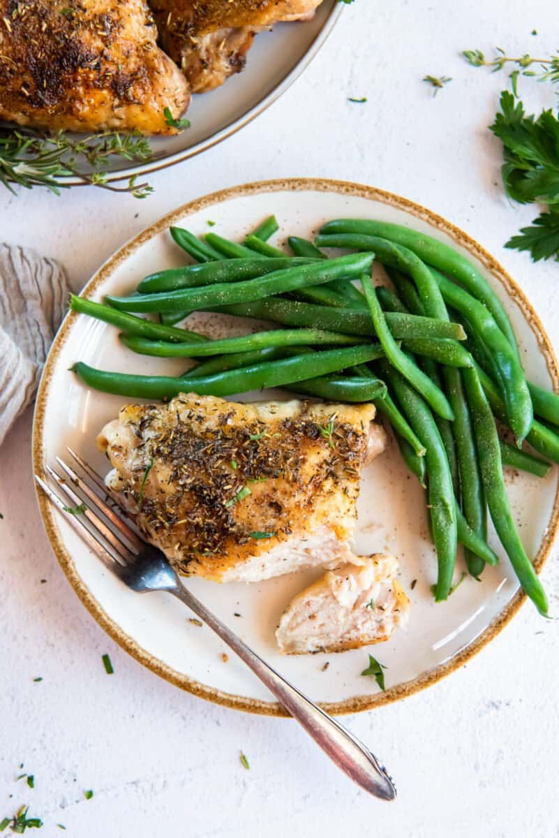 overhead image of baked chicken thigh on a white plate with green beans and a fork