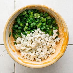blue cheese crumbles and sliced green onions added to buffalo cream cheese mixture in a bowl