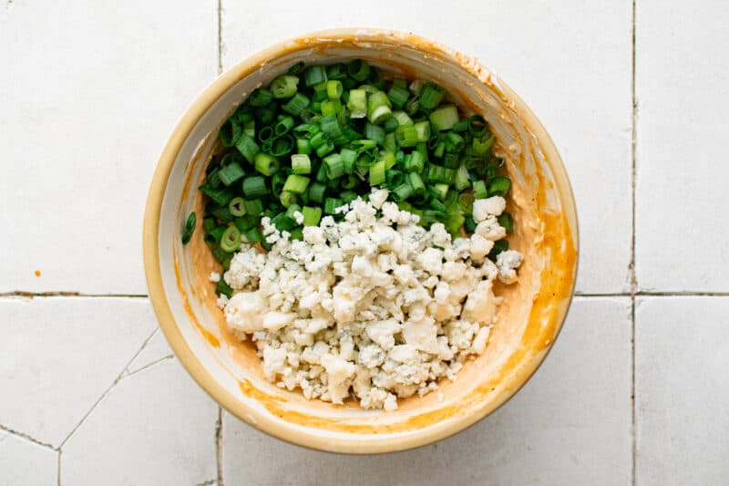 blue cheese crumbles and sliced green onions added to buffalo cream cheese mixture in a bowl