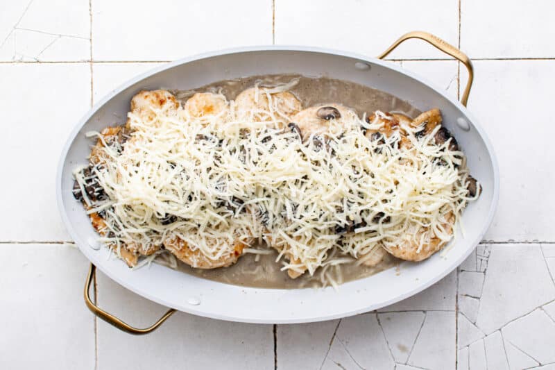 shredded cheese on top of chicken and mushroom sauce in a white baking dish before baking