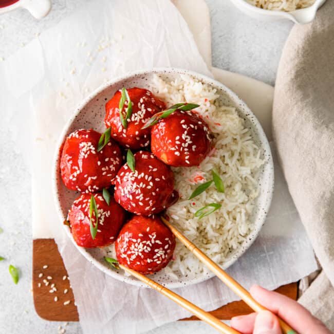 overhead image of sweet and sour chicken meatballs with rice on a white plate and a hand using chopsticks grabbing a meatball