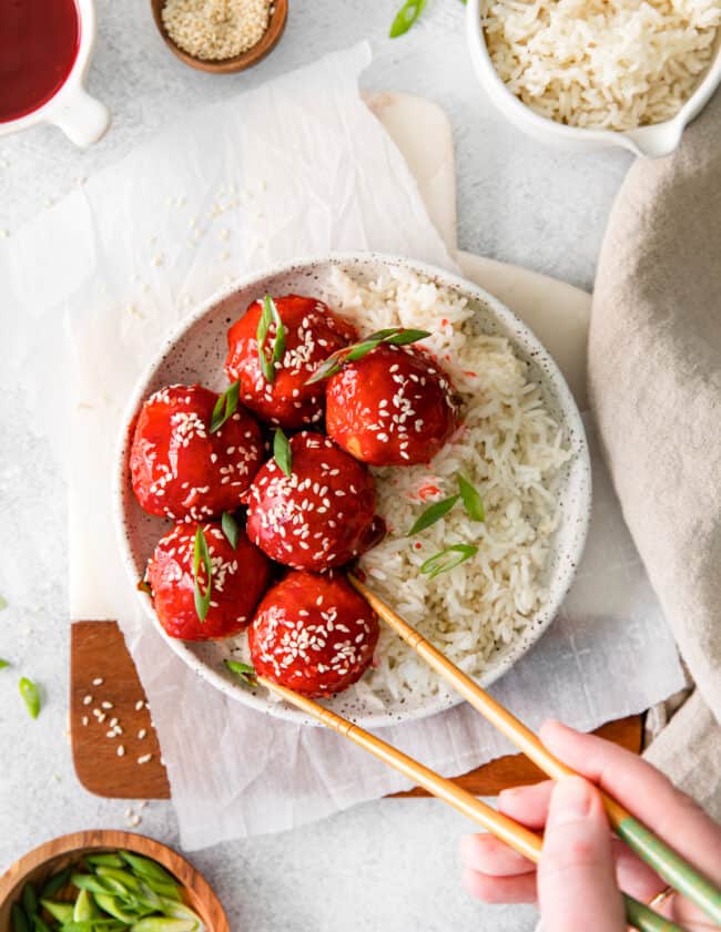 overhead image of sweet and sour chicken meatballs with rice on a white plate and a hand using chopsticks grabbing a meatball