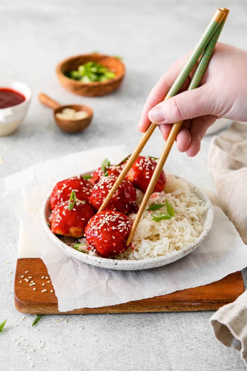 hand holding chopsticks grabbing a sweet and sour chicken meatball off of a white plate