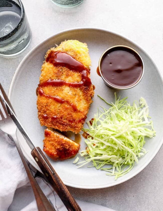 overhead view of chicken katsu with tonkatsu sauce and shredded cabbage on a white plate with a fork and knife.