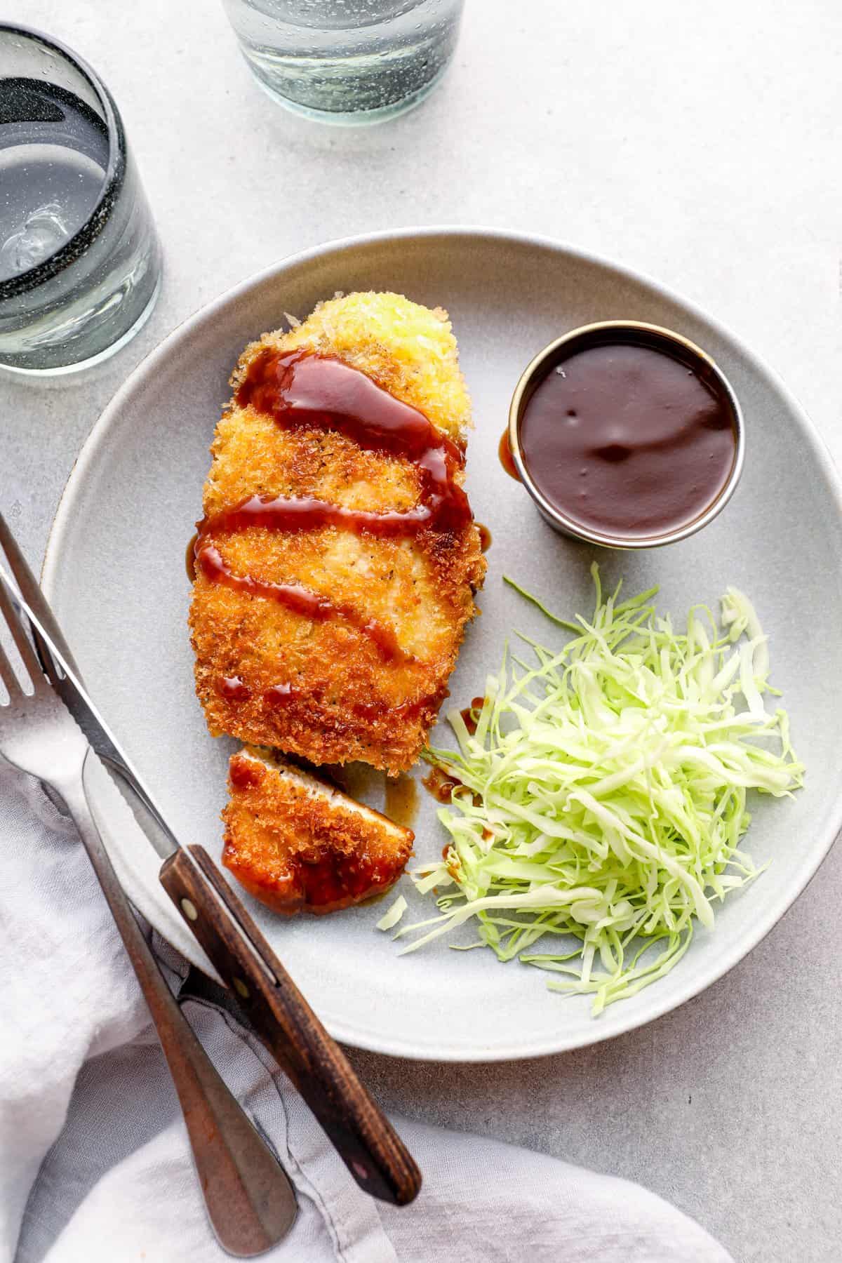 overhead view of chicken katsu with tonkatsu sauce and shredded cabbage on a white plate with a fork and knife.
