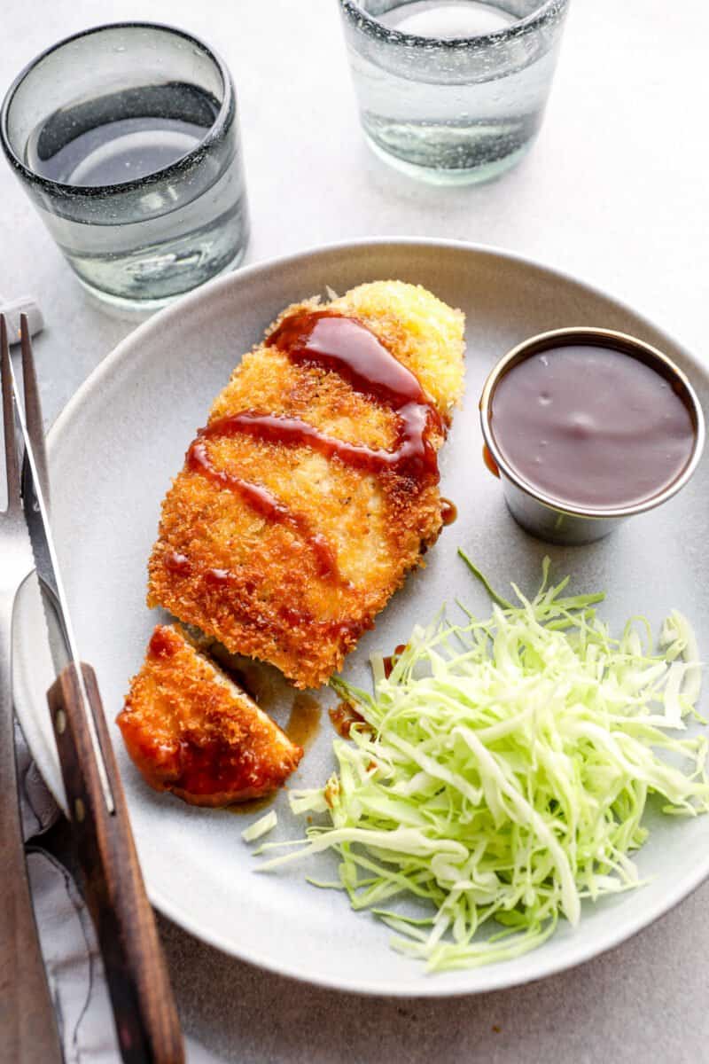 overhead view of cut chicken katsu with tonkatsu sauce and shredded cabbage on a white plate with a fork and knife.