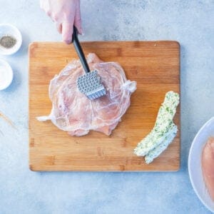 overhead view of chicken breast being pounded thin on a wood cutting board.