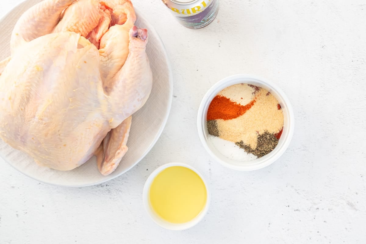 A raw whole chicken next to bowls with olive oil and beer can chicken seasoning.