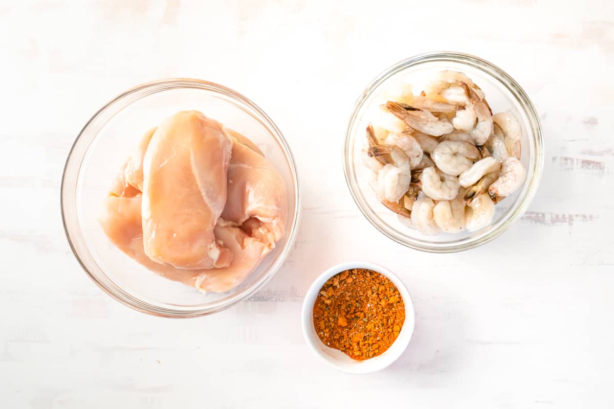Raw shrimp and raw chicken breasts in two separate glass bowls, next to a bowl of Cajun seasoning.