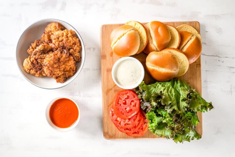 overhead view of fried buffalo chicken next to buns lettuce tomatoes buffalo sauce and bleu cheese.
