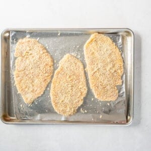 3 breaded chicken breasts on a baking sheet.