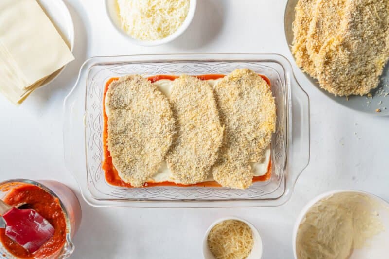 3 breaded chicken breasts on top of ricotta noodles and marinara in a glass baking dish.