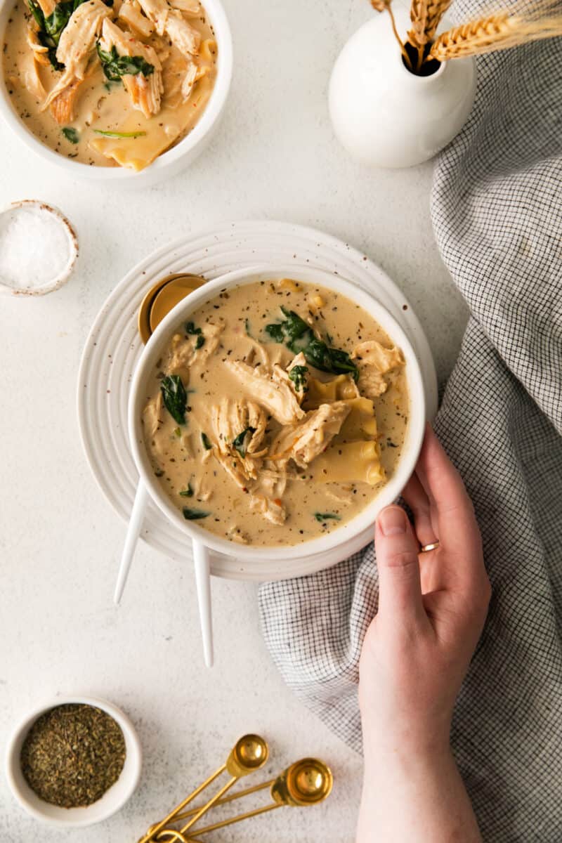 overhead view of a hand holding a bowl of white chicken lasagna soup on a white plate with spoons.