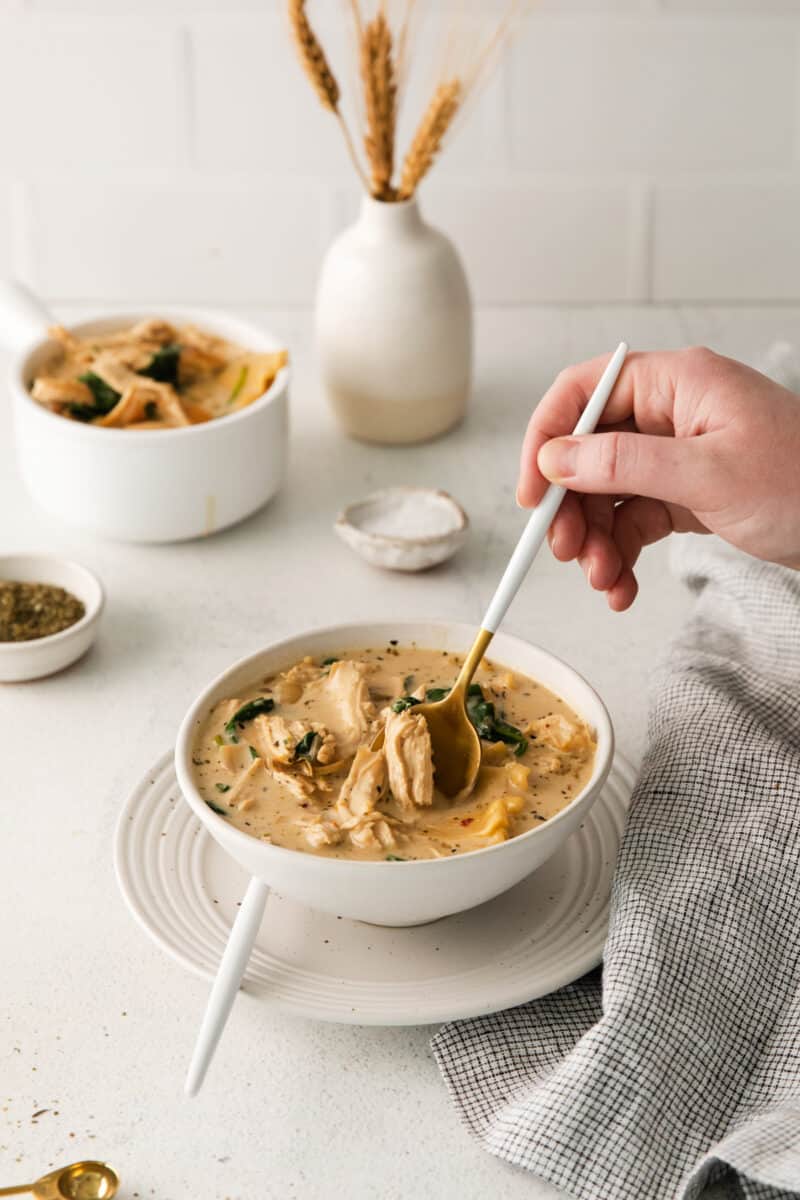 a hand scooping white chicken lasagna soup in a white bowl on a white plate with spoons.