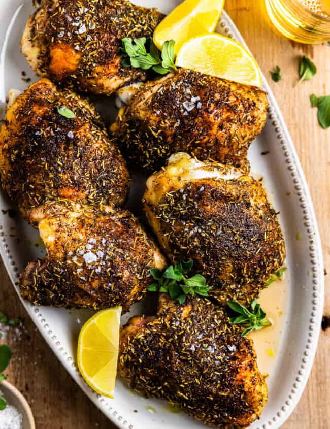 overhead view of baked chicken thighs on a white oval platter with lemon wedges.