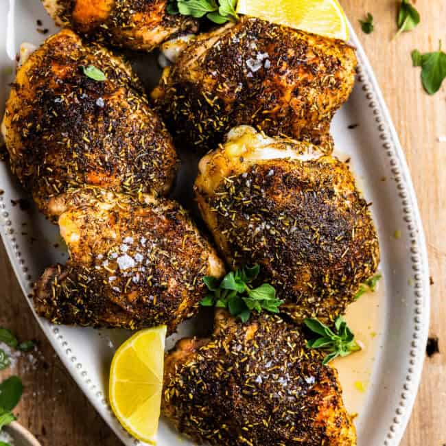 overhead view of baked chicken thighs on a white oval platter with lemon wedges.