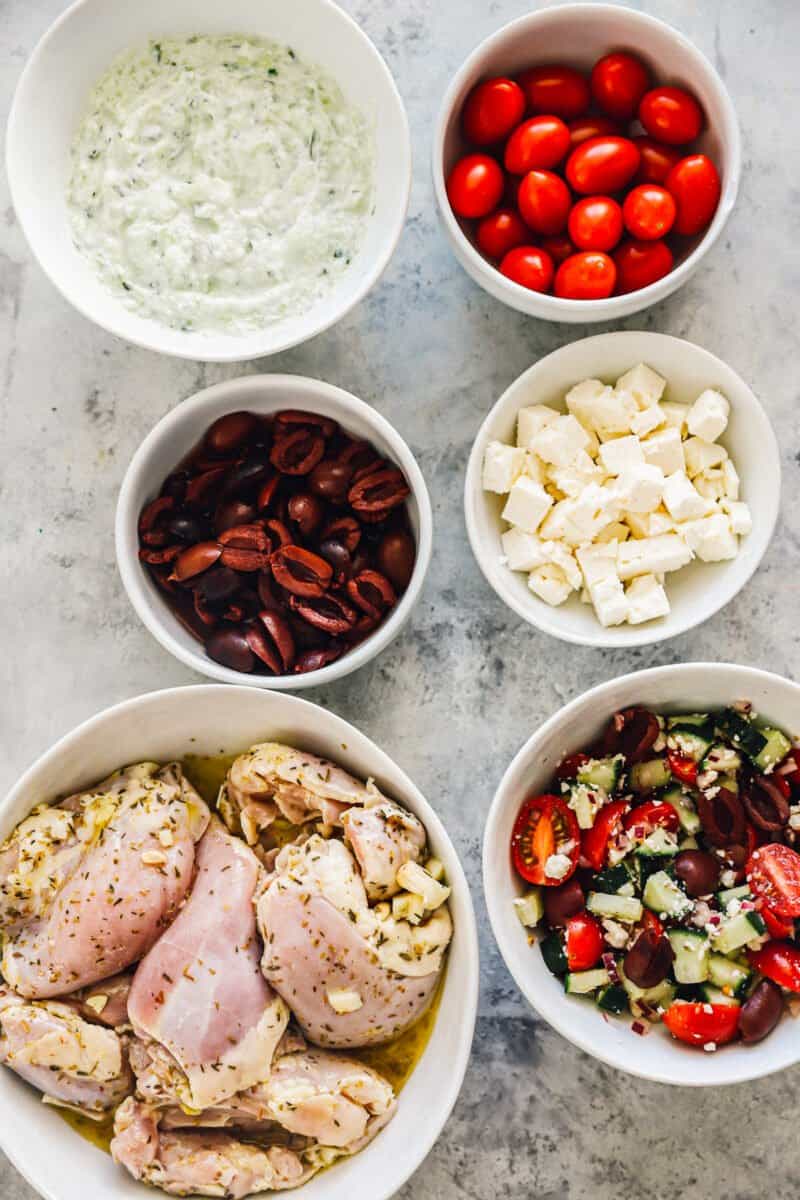 bowls of tomatoes, olives, feta, tzatziki, greek salad, and chicken thighs arranged on a table top