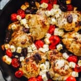 greek chicken thighs in a cast iron skillet, surrounded by tomatoes and feta