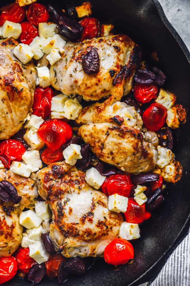 a skillet filled with chicken thighs, tomatoes, olives, and feta