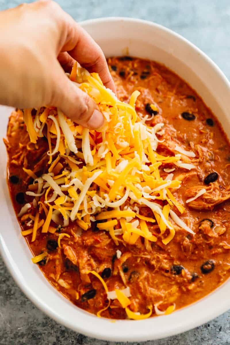 a hand placing shredded cheese into a baking dish filled with enchilada dip ingredients