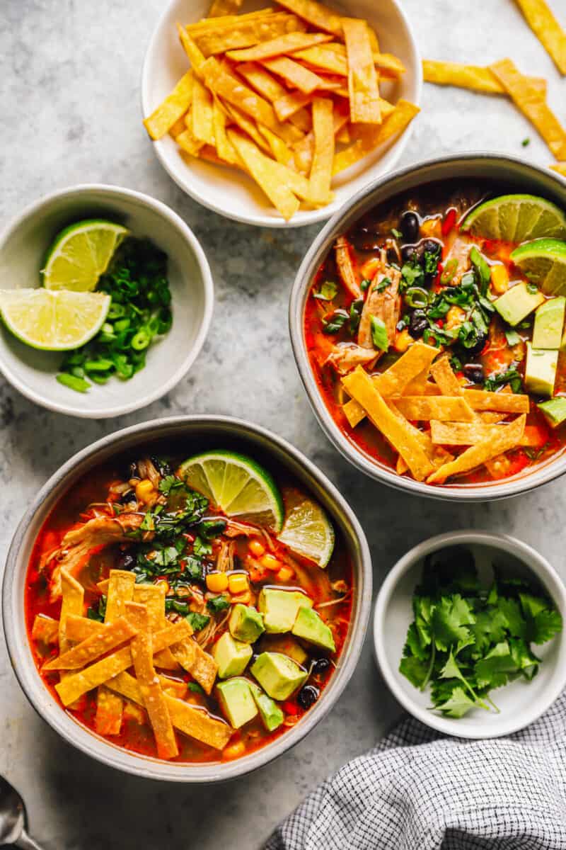 two bowls of easy chicken tortilla soup next to bowls filled with tortilla strips, limes, and cilantro