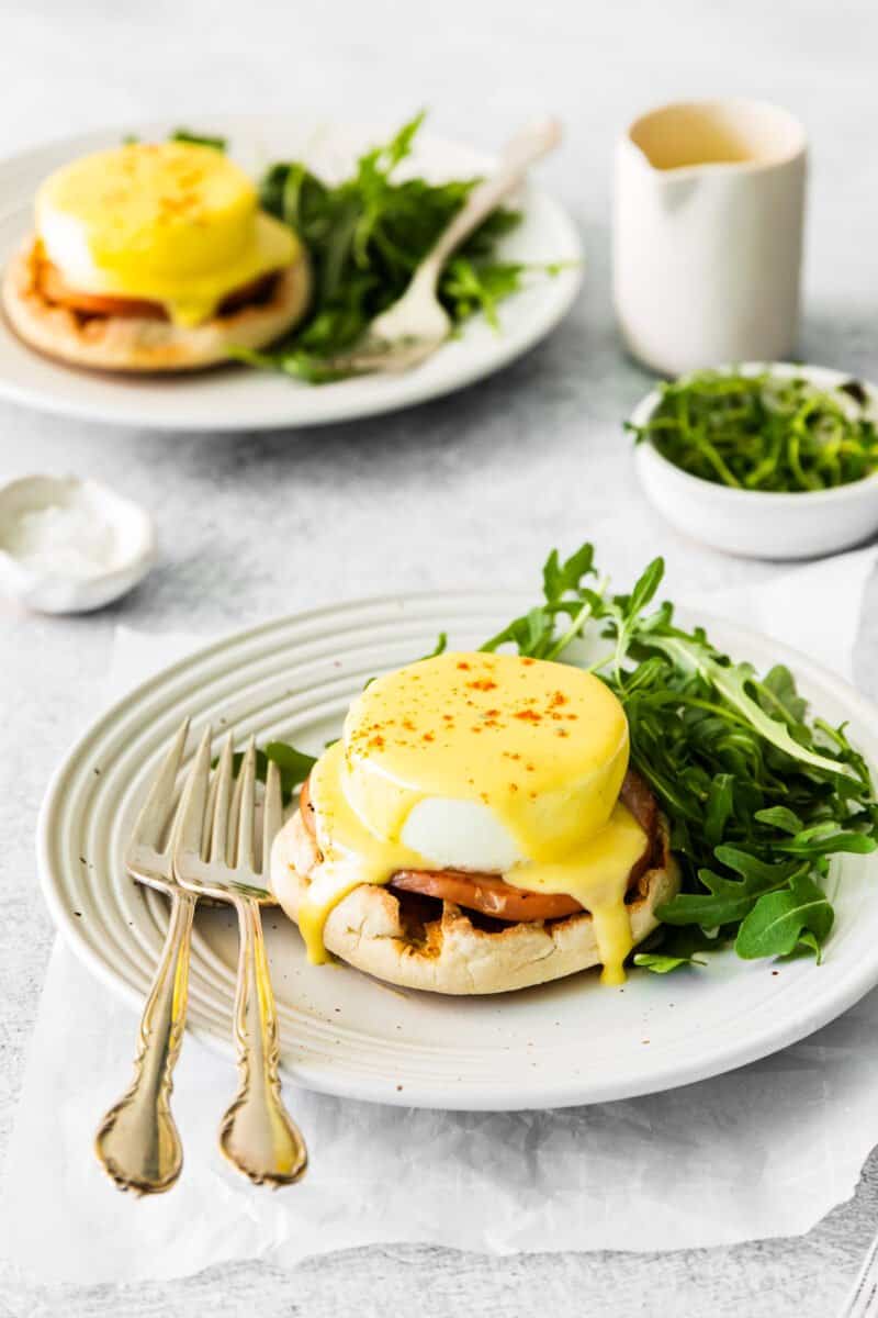 eggs benedict on a white plate with arugula and forks.