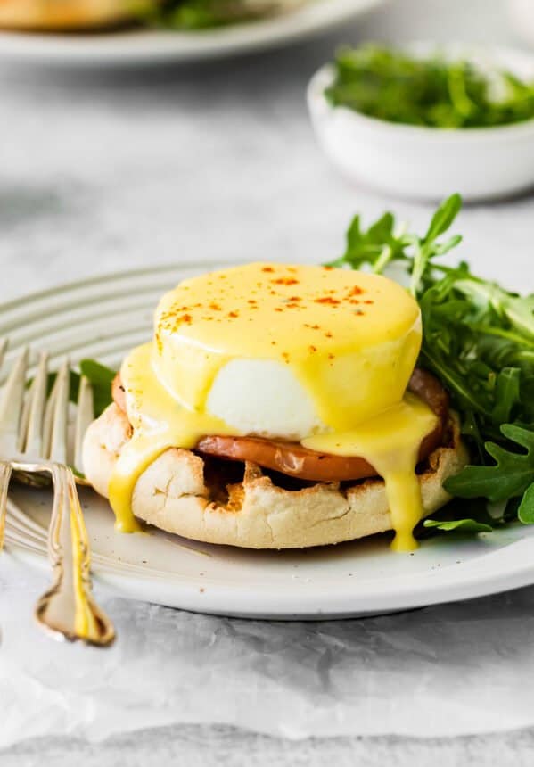 eggs benedict on a white plate with arugula and a fork.