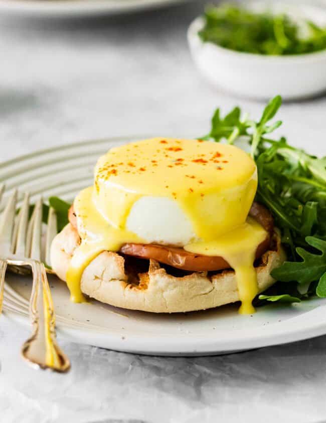 eggs benedict on a white plate with arugula and a fork.