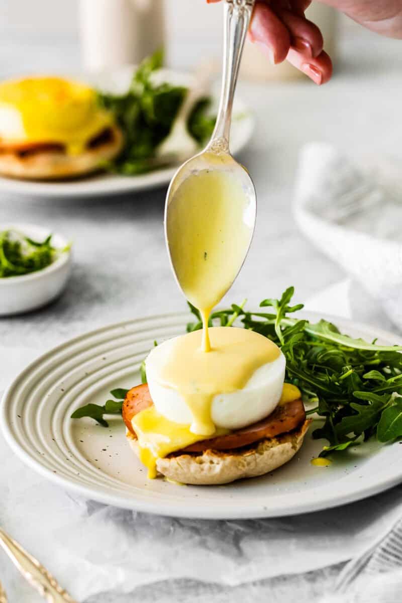 a spoon drizzling hollandaise sauce on eggs benedict on a white plate with arugula.