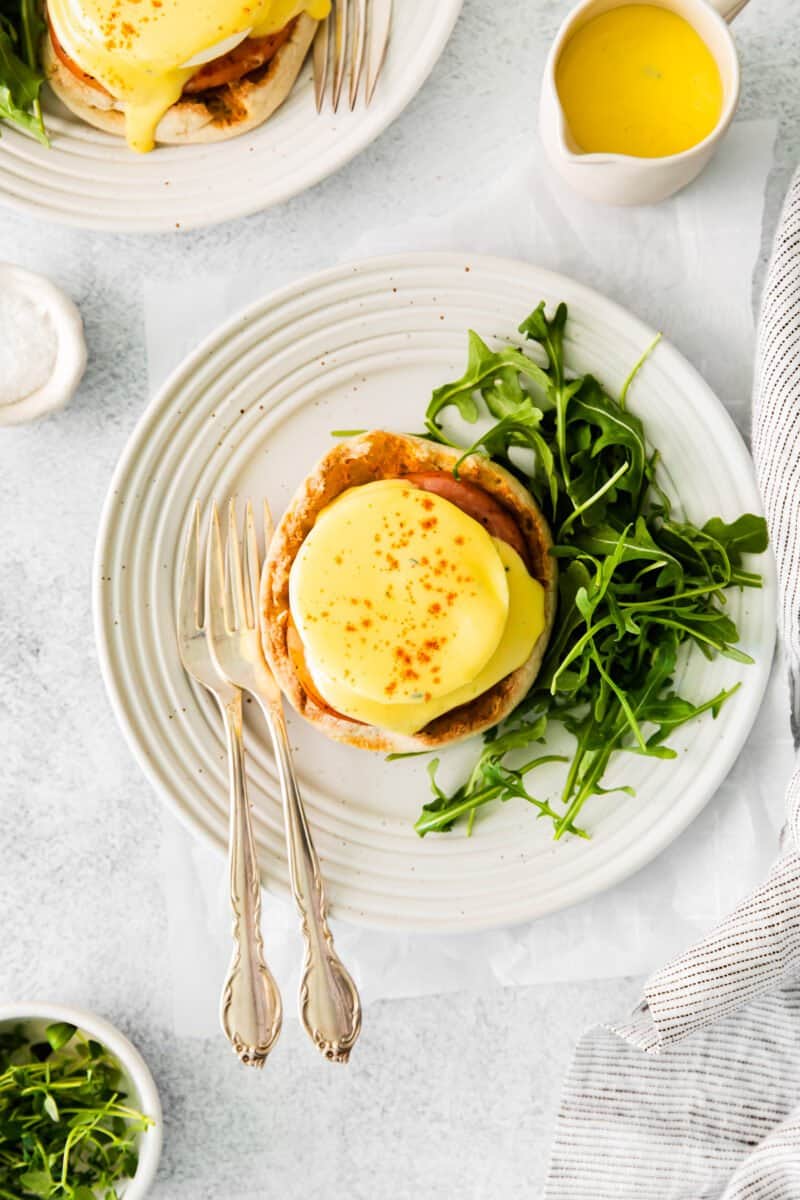 overhead view of eggs benedict on a white plate with arugula and forks.