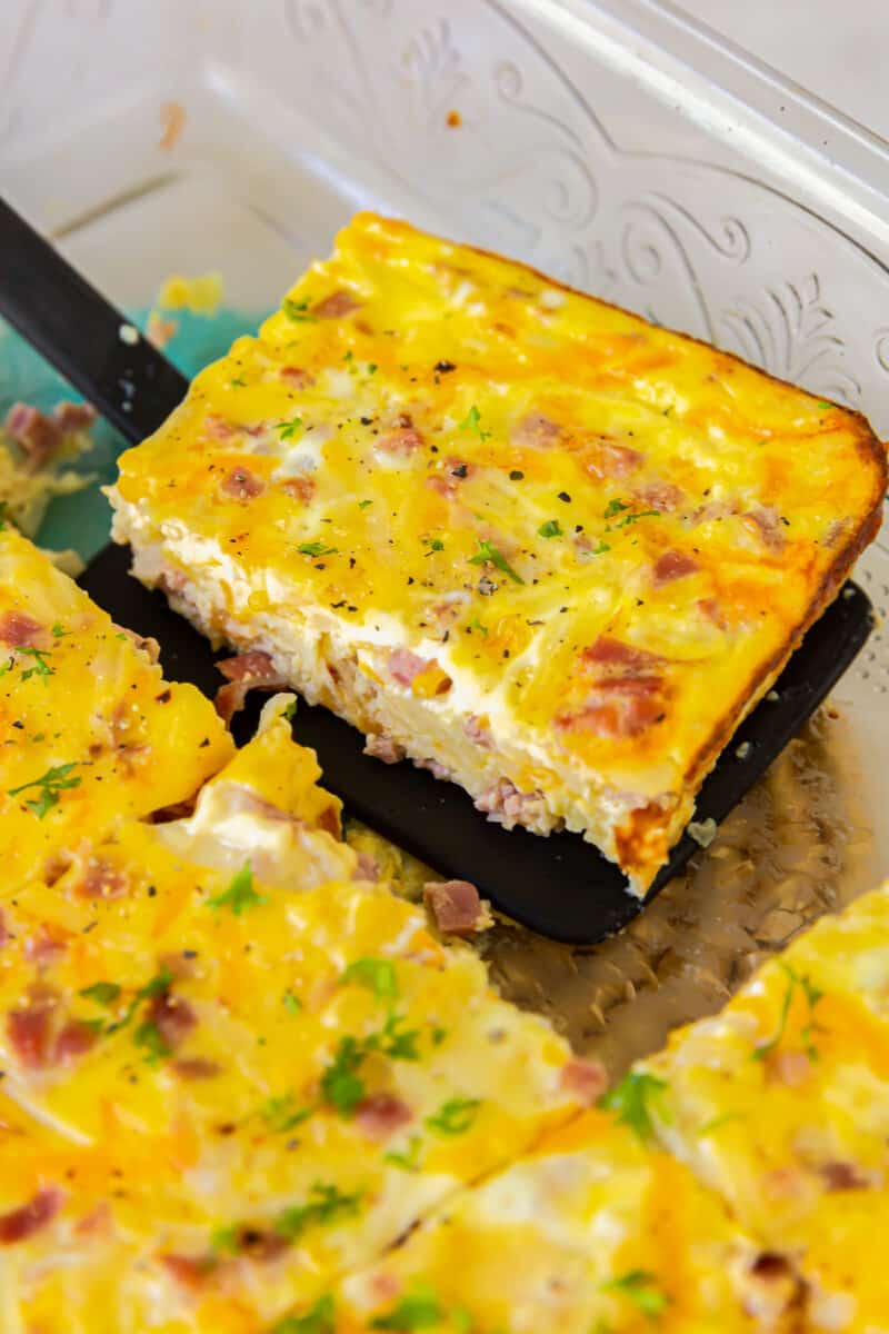 a spatula lifting a slice of ham and cheese breakfast casserole from a casserole dish.