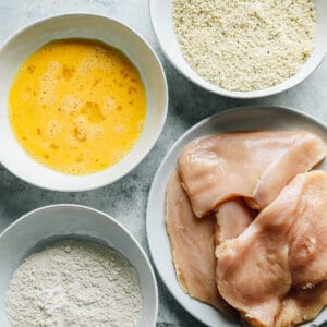 chicken dredging ingredients in white dishes (egg, flour, breadcrumbs, raw chicken)