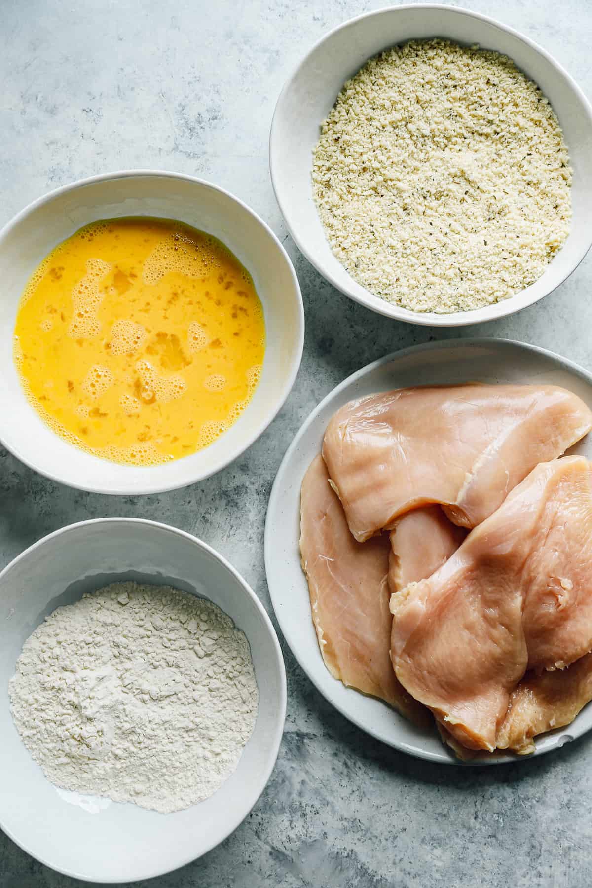 chicken dredging ingredients in white dishes (egg, flour, breadcrumbs, raw chicken)