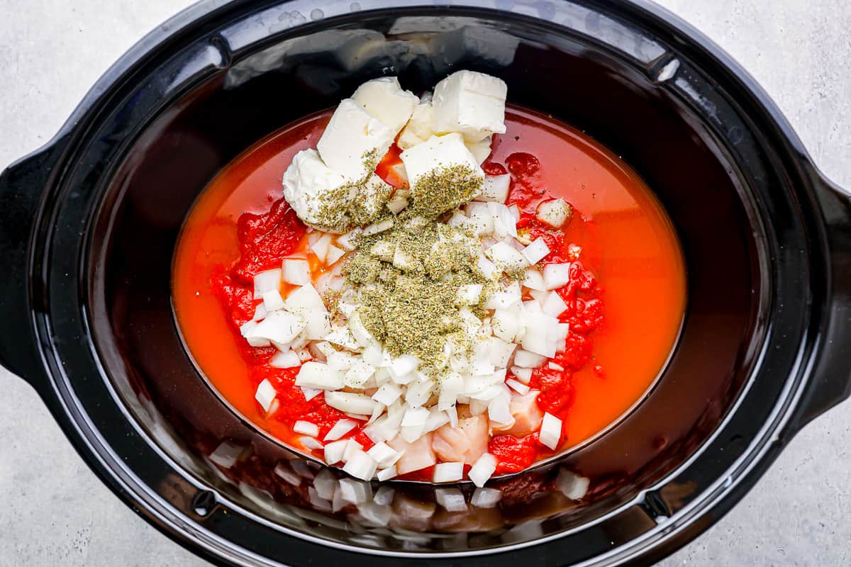 ingredients for crockpot chicken spaghetti in a slow cooker: cubes of cream cheese, marinara sauce, diced onions, seasonings, and chicken breast.