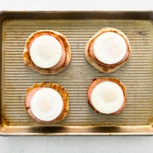 4 english muffins topped with canadian bacon and poached eggs on a baking sheet.