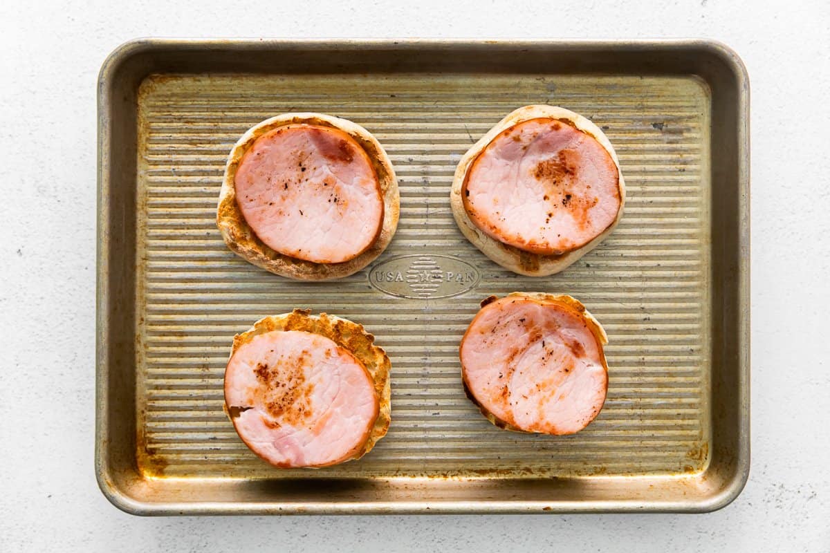 4 english muffins topped with canadian bacon on a baking sheet.